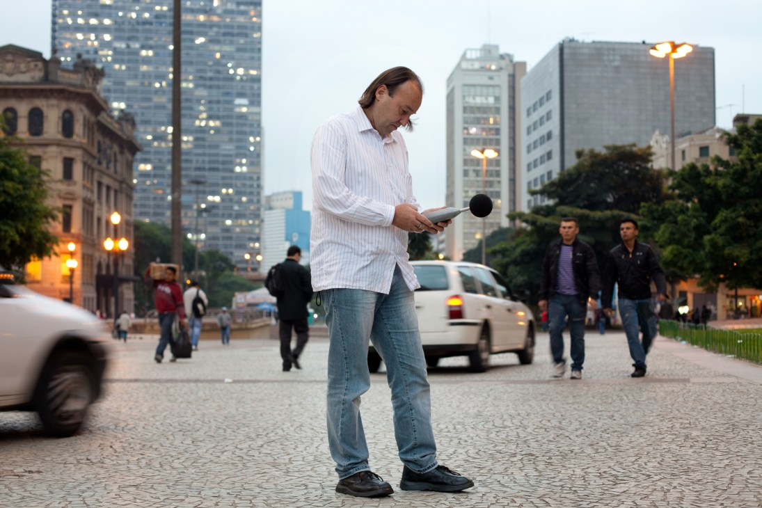 Man in white shirt holding decibel meter on São Paulo street surrounded by buildings and pedestrians.