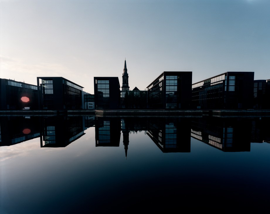 Modern black buildings reflected in still water with a church spire visible in the background