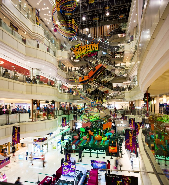 Interior of Grand Shopping Centre showing multiple levels with escalators, storefronts, and colorful promotional displays.