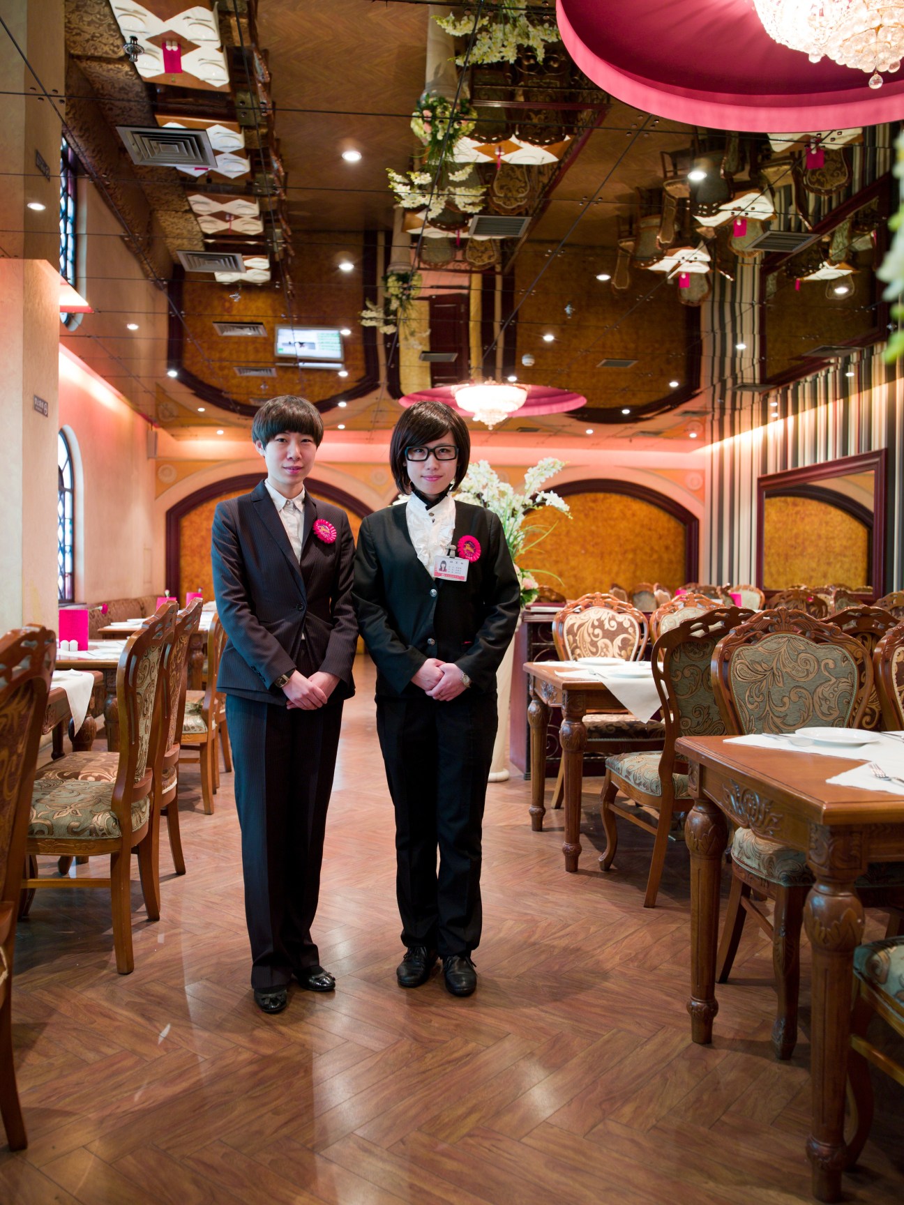 Two uniformed staff members stand in an elegant restaurant dining room with ornate wooden chairs and pink decorative elements.