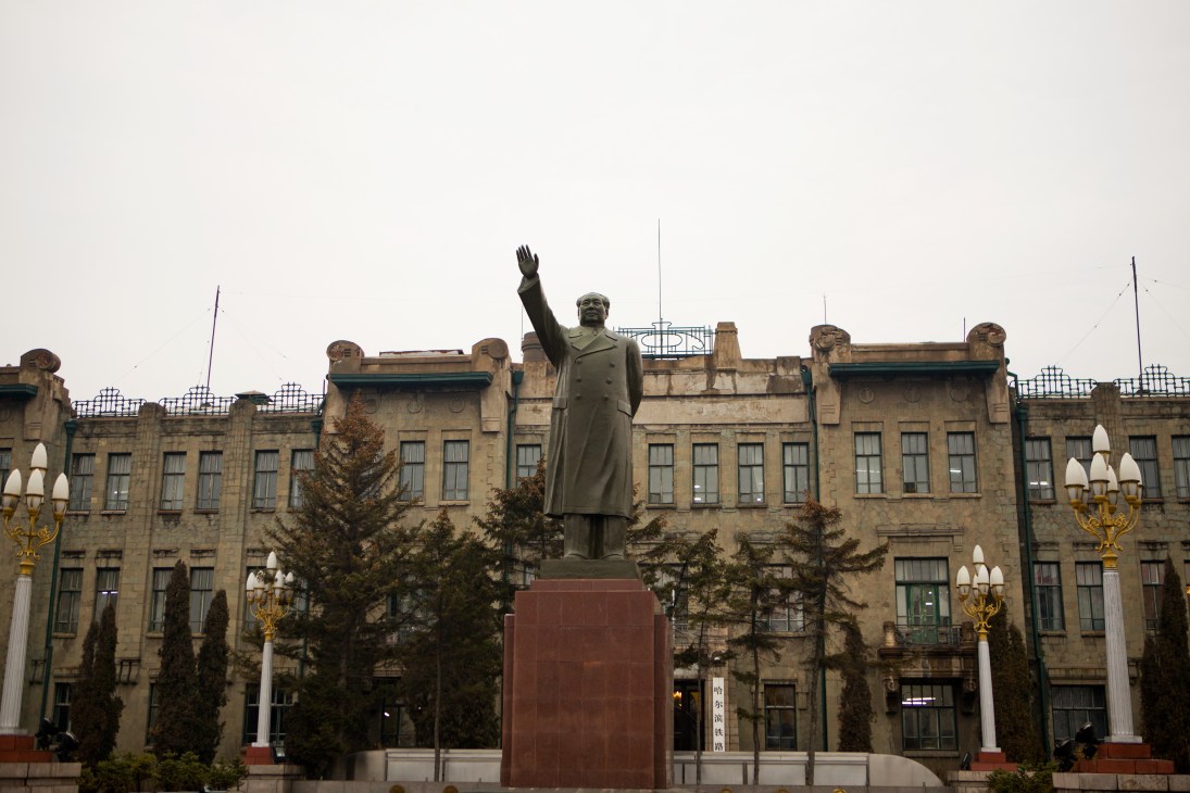 Statue of Mao Zedong with raised arm on red pedestal in front of stone building.