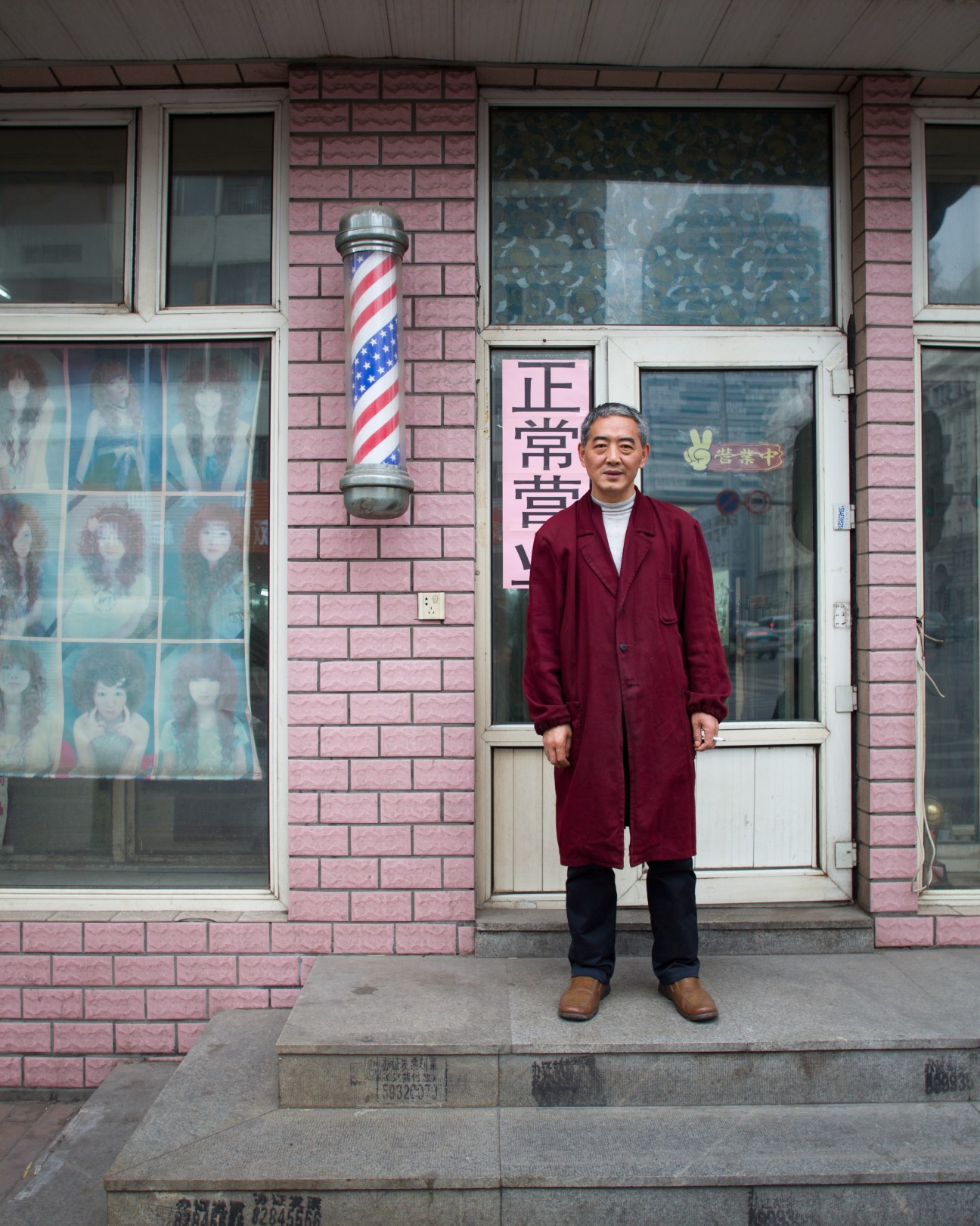 Man in maroon coat stands outside a pink brick barber shop with red and white striped pole and Chinese signage.