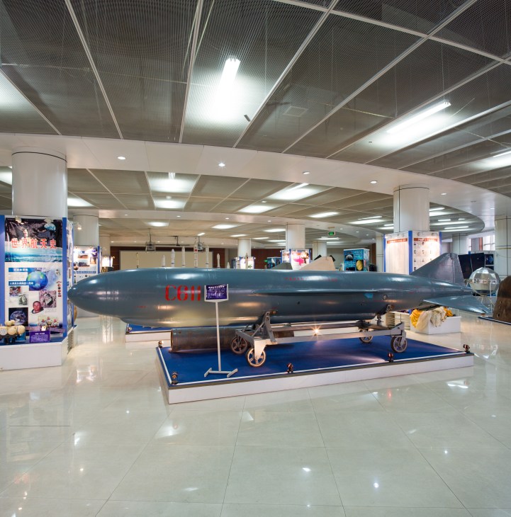 A large dark blue missile displayed on a blue platform inside a modern museum with white columns and fluorescent ceiling lights.
