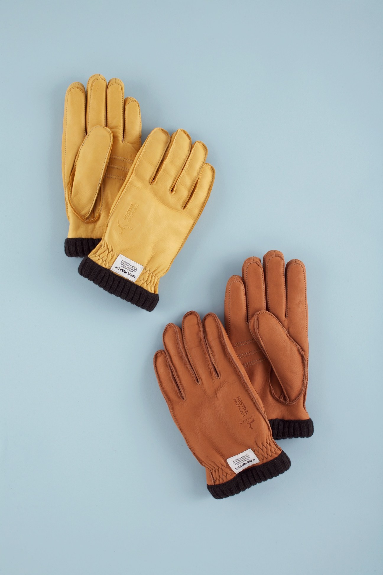 Two pairs of deerskin gloves in yellow and tan with dark ribbed cuffs on a light blue background.