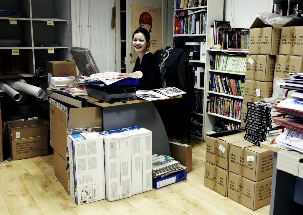 Editorial assistant Lizzie Kaye smiles while working at a desk surrounded by boxes of graphic novels and publishing materials.