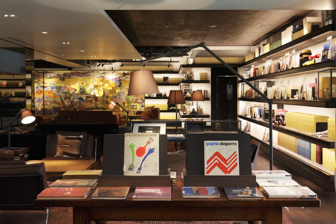Interior of Tsutaya Books' Anjin lounge with armchairs, pendant lamps, artwork, shelving, and graphic design posters on display.