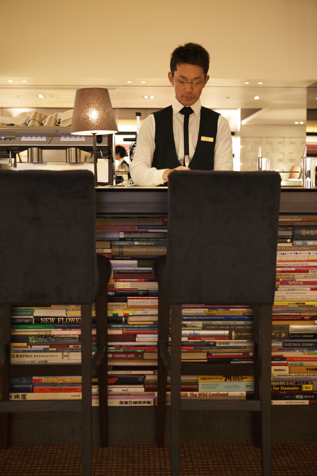 A concierge staff member in formal white shirt and black vest stands behind a counter in Tsutaya Books' lounge area.