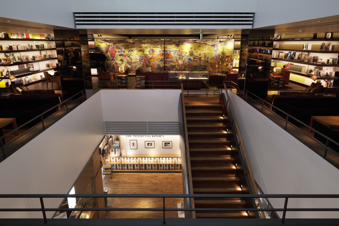 Interior of Tsutaya Books showing stairway with illuminated steps, bookshelves lining upper level, and colorful artwork on back wall.