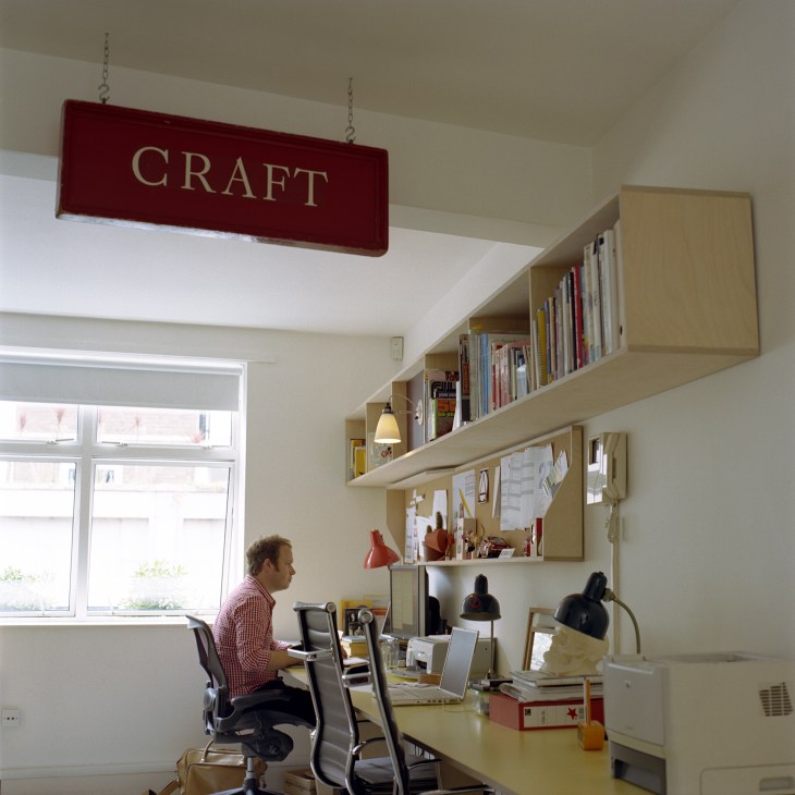 Designer working at desk in bright office with shelving and 'CRAFT' sign overhead.