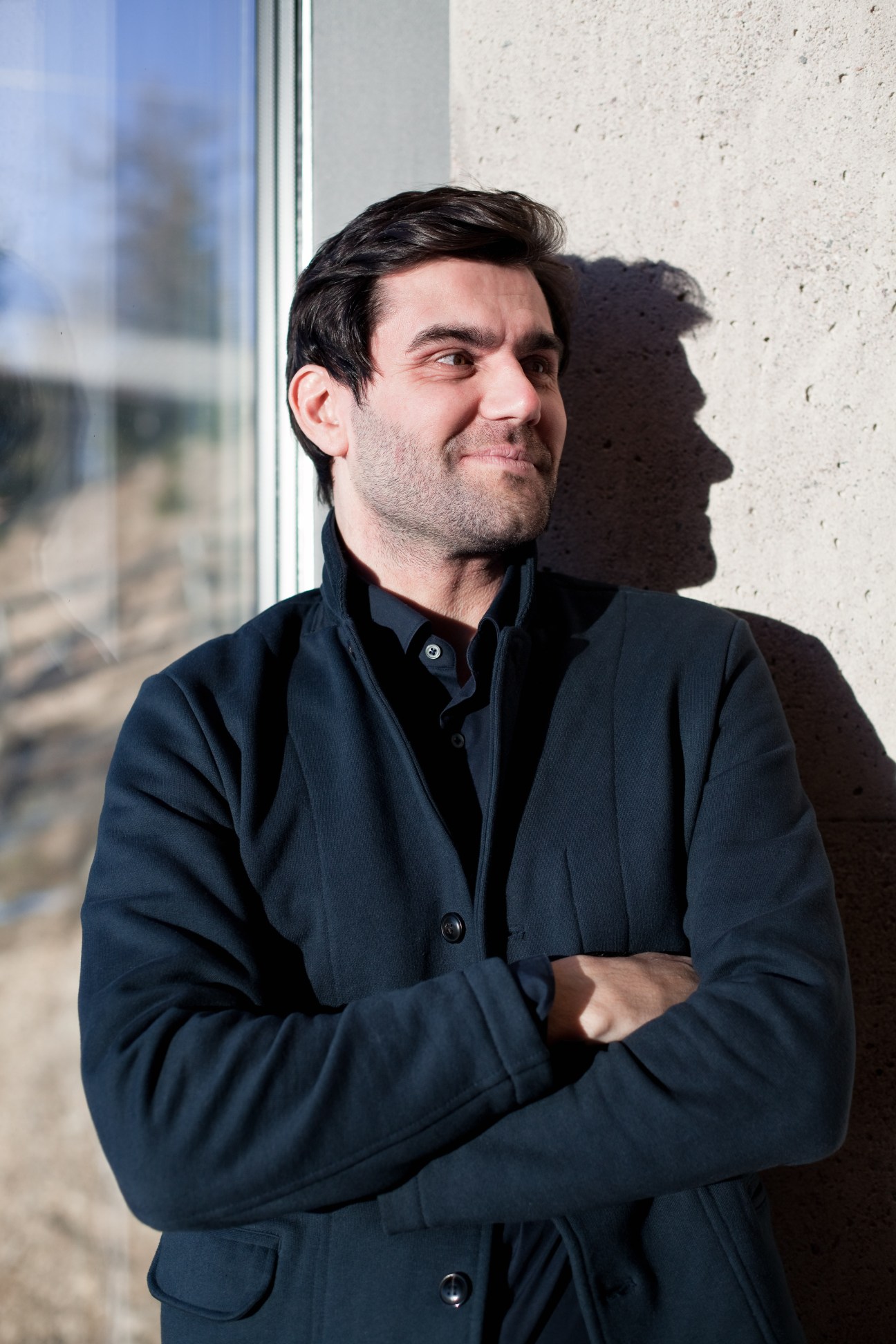Architect Peter Pichler stands by a window with a concrete wall behind him, arms crossed, looking to the side.