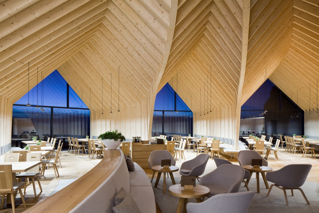 Interior of Oberholz mountain hut showing exposed spruce wood frame, pitched ceilings, dining tables with oak chairs, and floor-to-ceiling windows overlooking mountains.
