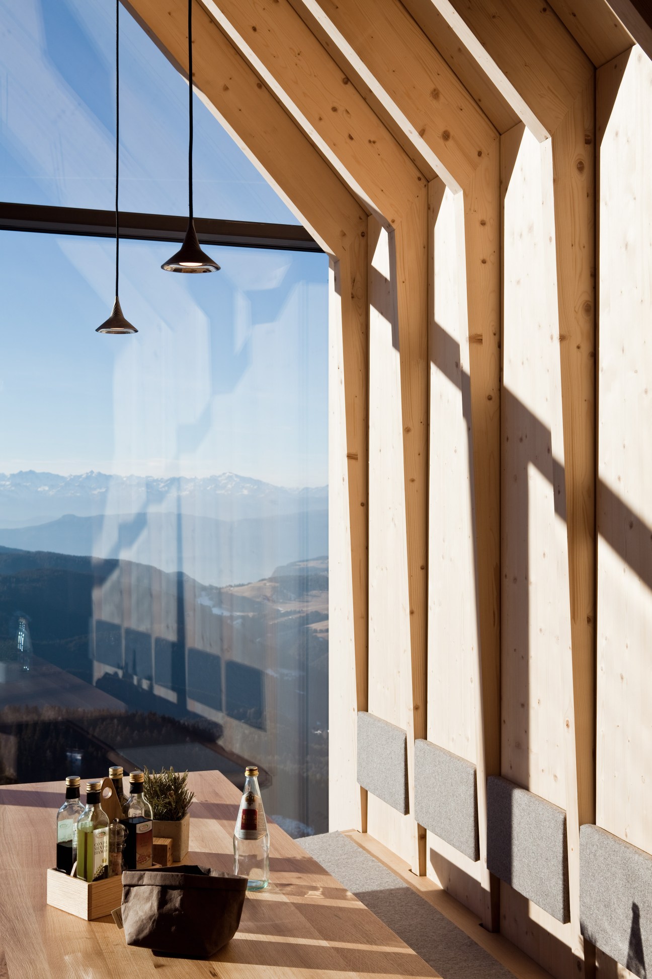 Oberholz mountain hut interior with large glass windows framing Alpine mountain views, wooden beams, pendant lamps, and a table with beverages.