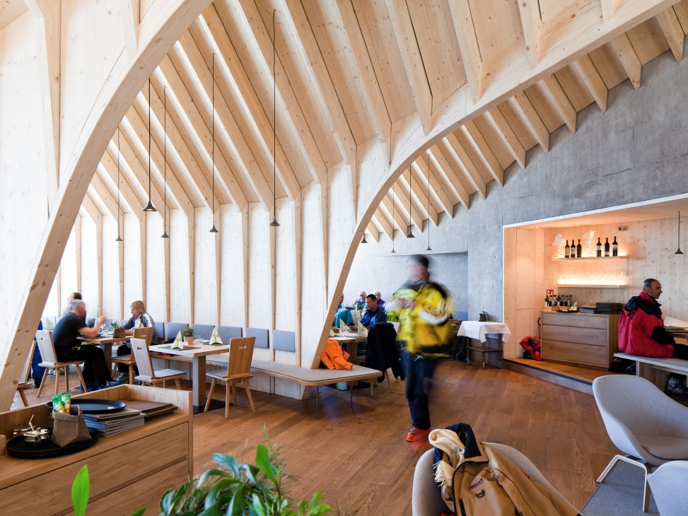 Interior of Oberholz mountain hut with curved spruce wood frame, diners at oak tables, and bar area with bottle display.
