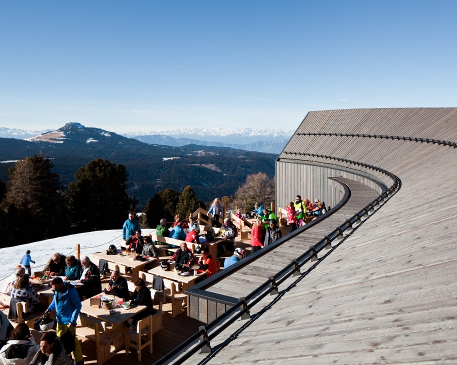 Diners enjoy lunch on the Oberholz alpine hut's curved wooden terrace with panoramic Dolomite mountain views.