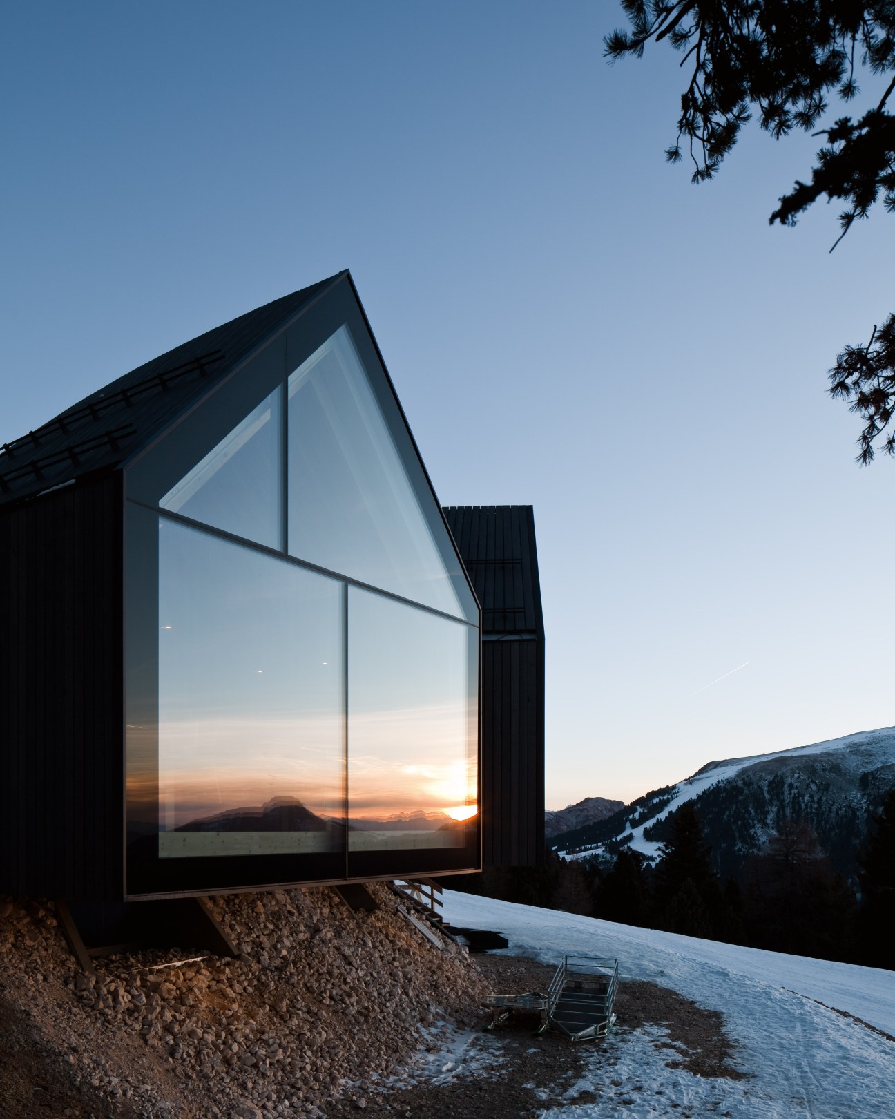 Oberholz alpine hut exterior with large triangular glass windows framing a mountain sunset view, dark wood cladding, and snowy landscape.