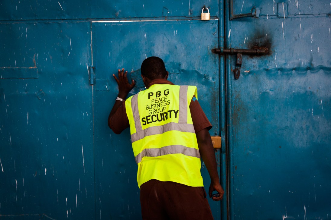 Security guard in high-visibility vest looking through hole in blue gate at Peace Hotel in Mogadishu