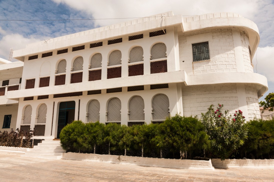 White curved building with mesh-covered arched windows and green shrubs at ground level.