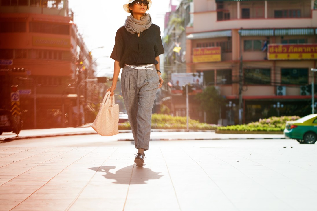 Woman in neutral tones walking through Bangkok street with sunglasses and canvas bag.