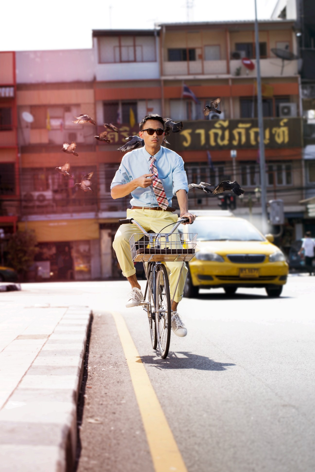 Man on bicycle riding through Bangkok street wearing light blue shirt and yellow trousers with birds flying nearby.