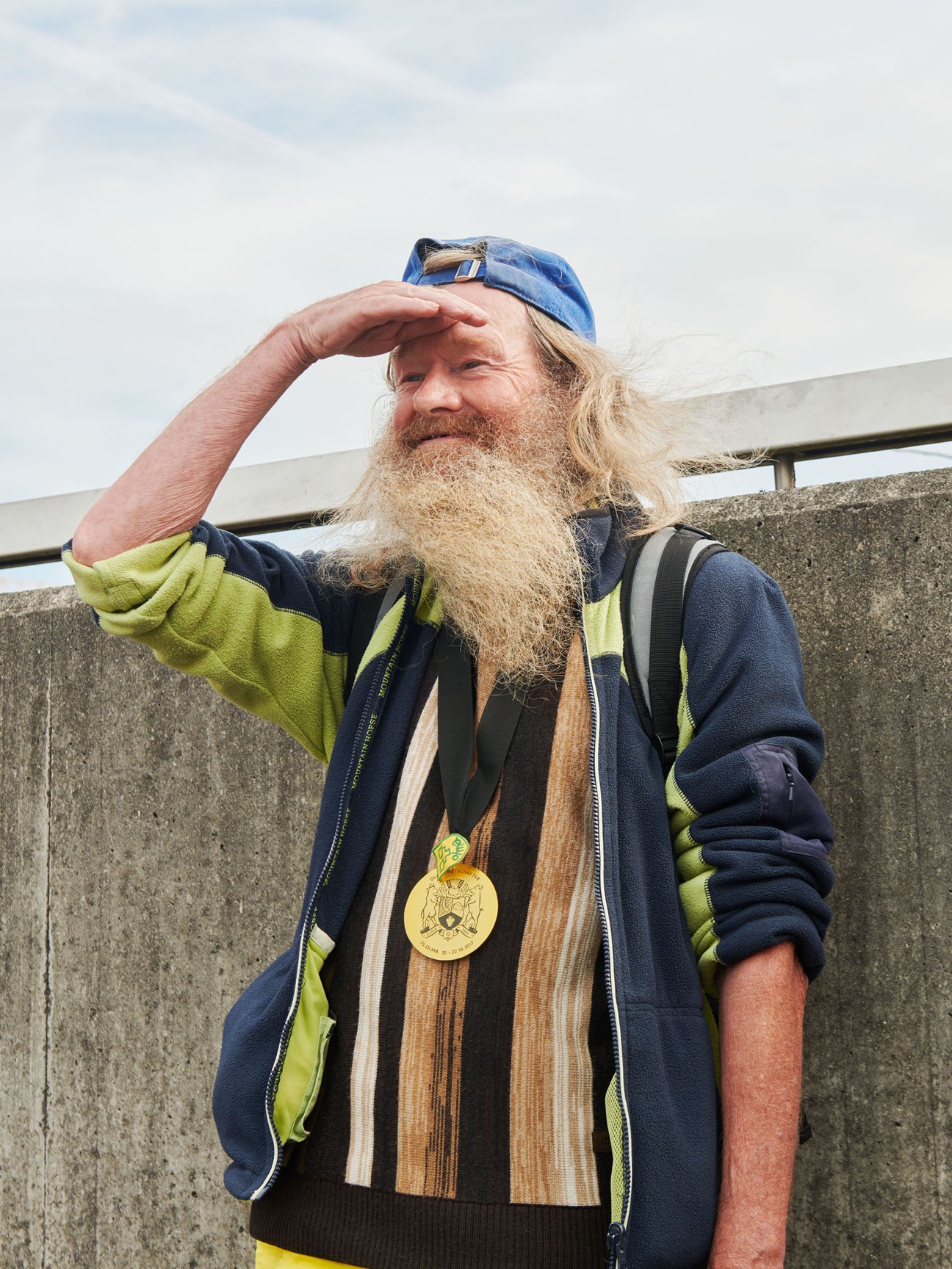Man with long grey beard wearing blue cap and colourful jacket holds hand to forehead, gold medal visible around his neck.