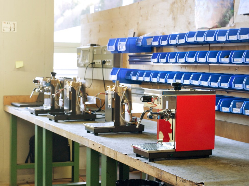 Red Olympia espresso machine on factory workbench with brass testing equipment and blue storage bins in background