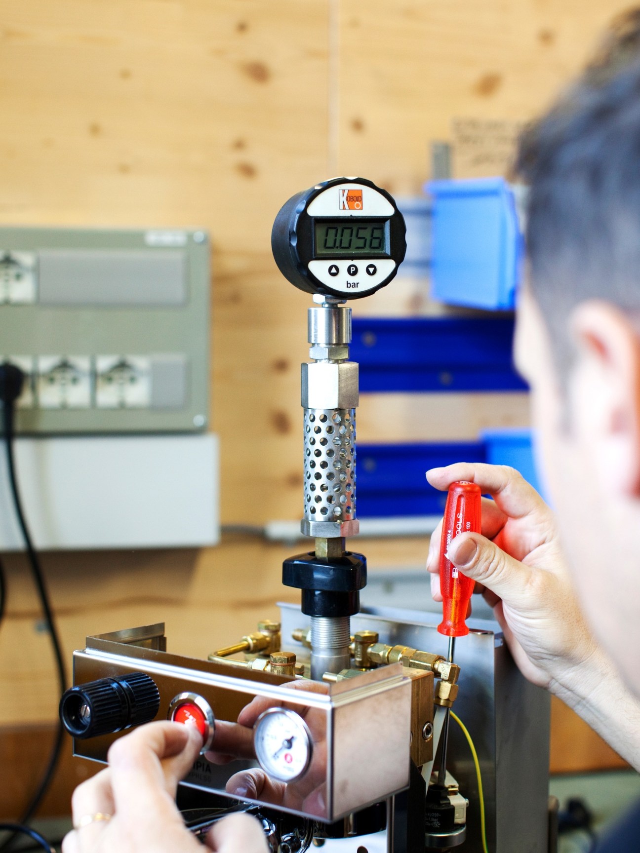 Worker measures pressure reading on digital gauge during Olympia espresso machine assembly.
