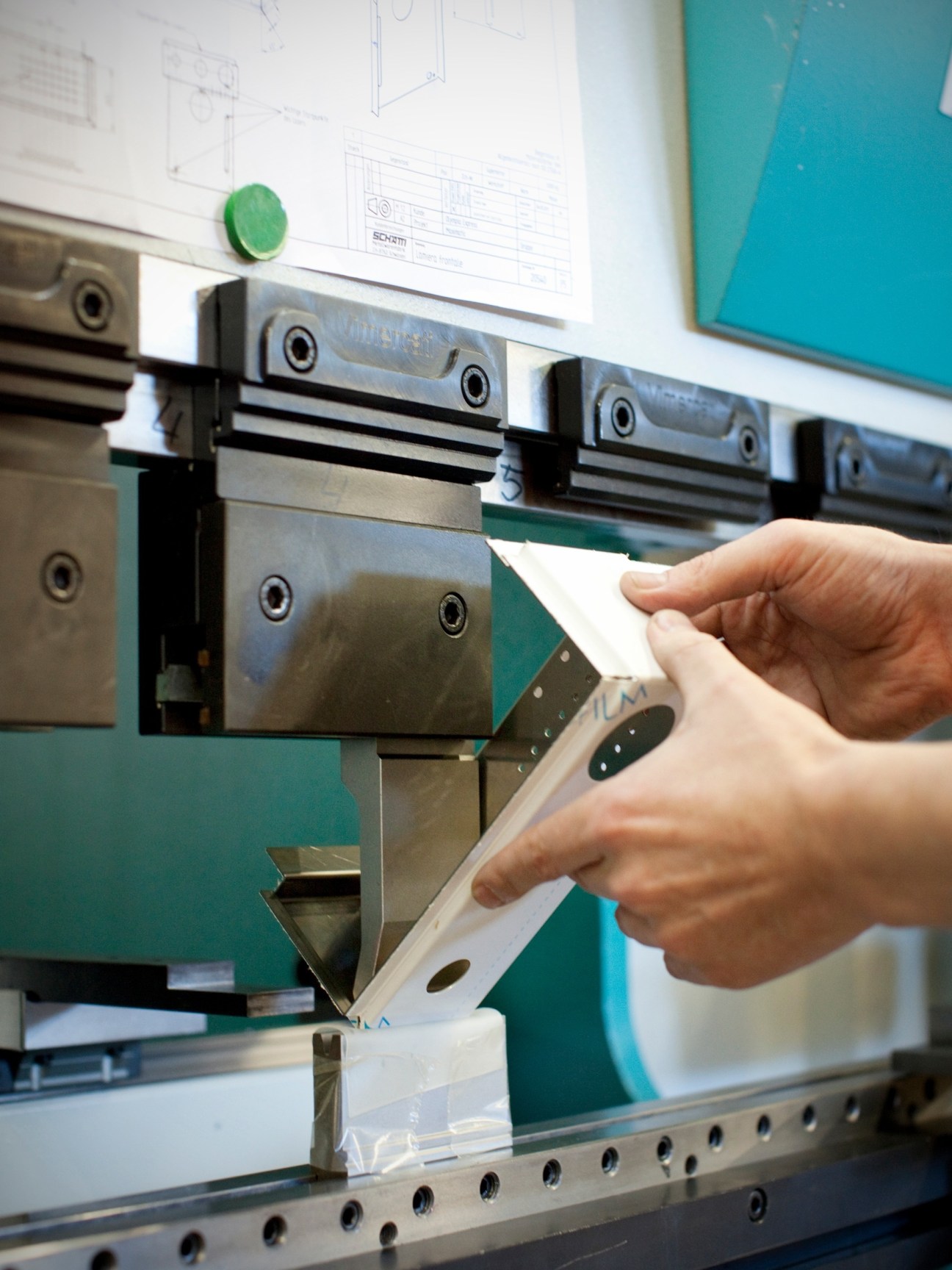 Worker's hands feeding a metal sheet into a green industrial bending machine with technical drawings visible on the wall behind.