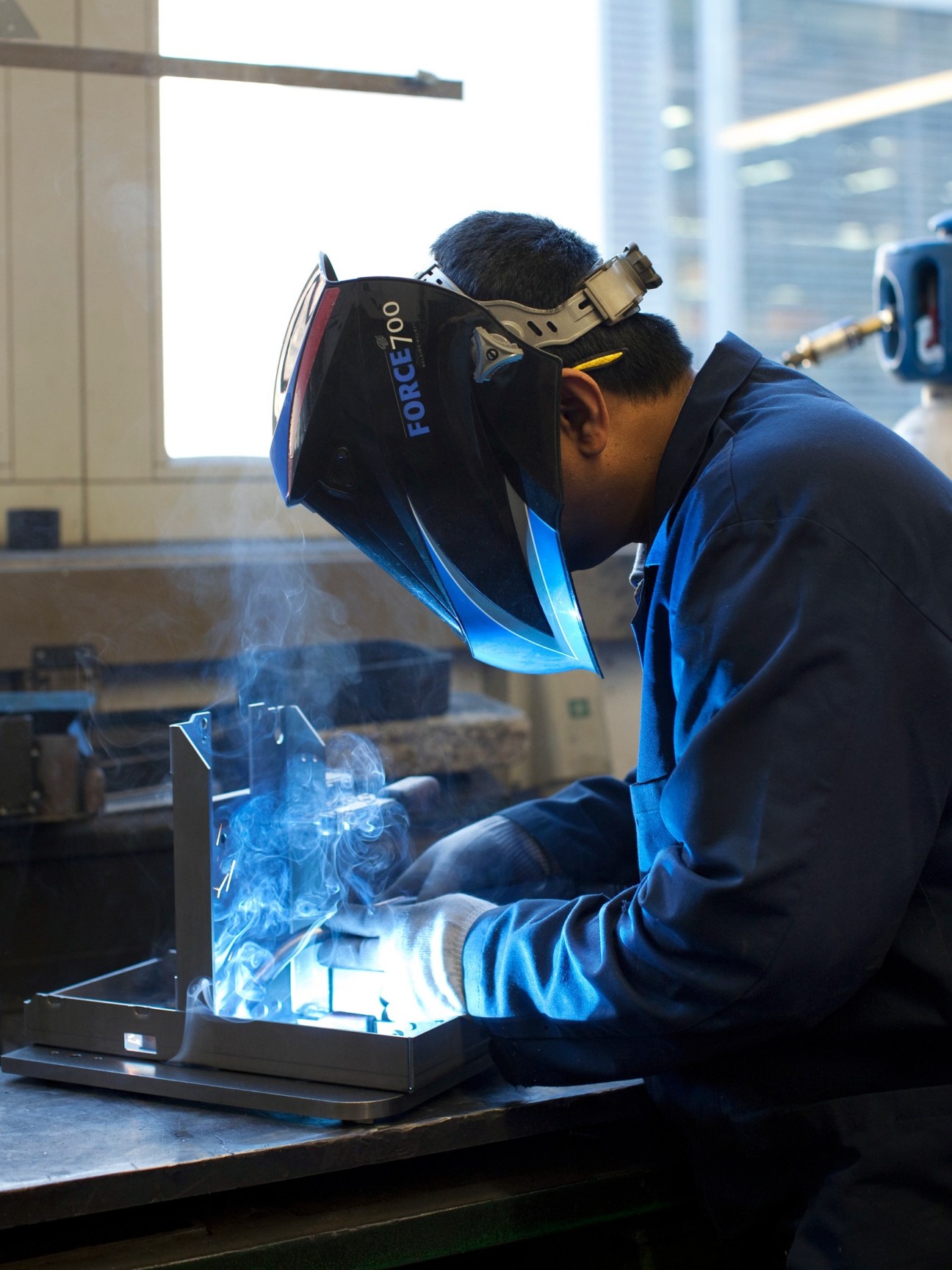 Worker in blue uniform and protective welding helmet welding metal parts for an espresso machine.