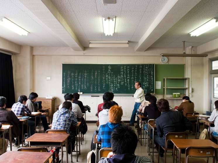 A classroom in Japan with students seated at desks facing a teacher standing near a chalkboard covered in Japanese characters.