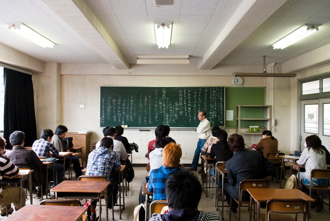 A classroom in Japan with students seated at desks facing a teacher standing near a chalkboard covered in Japanese characters.