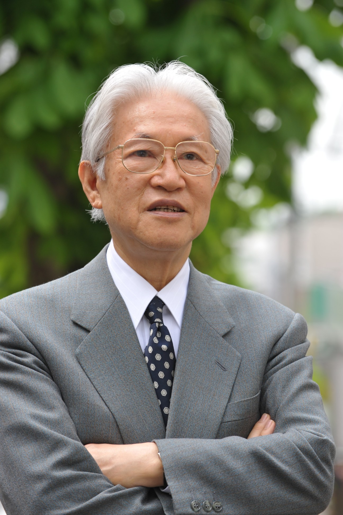 Elderly Asian man with white hair wearing glasses, gray suit and patterned tie, standing outdoors with green foliage background.