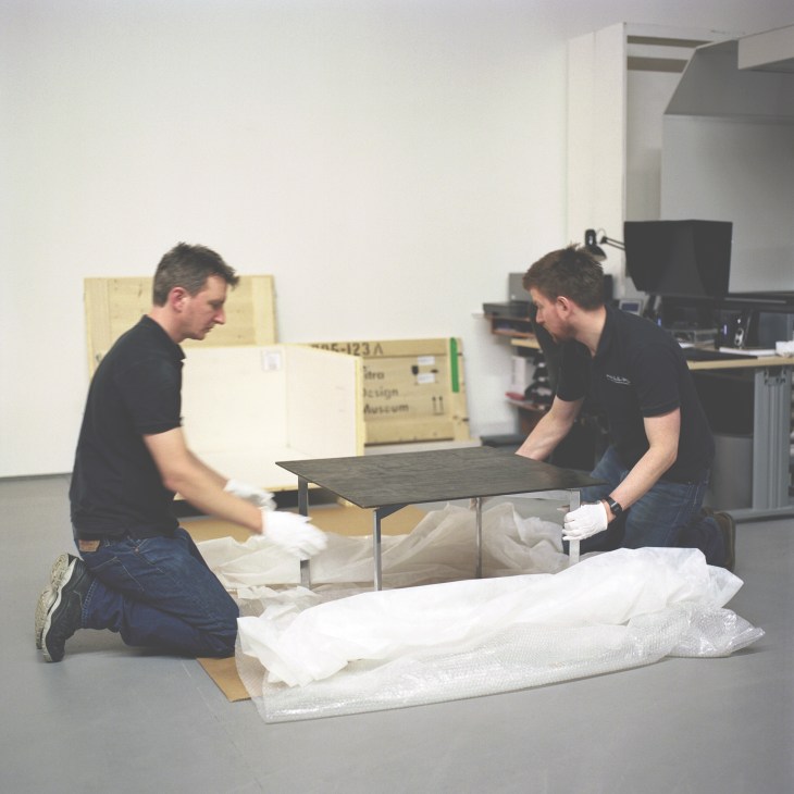 Two staff members in black shirts carefully unpack a wrapped design table in a white studio space.