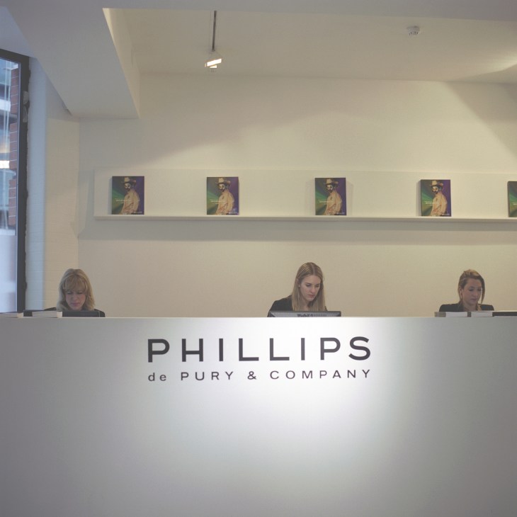 Phillips de Pury & Company reception desk with three staff members seated behind it and artwork displayed on wall above.
