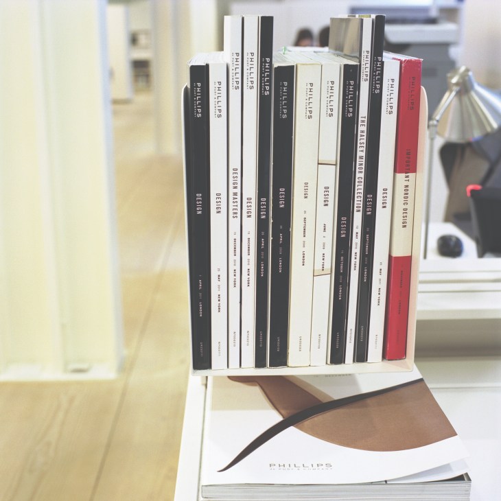 Phillips de Pury design auction catalogues with black, white and red spines displayed on a shelf.