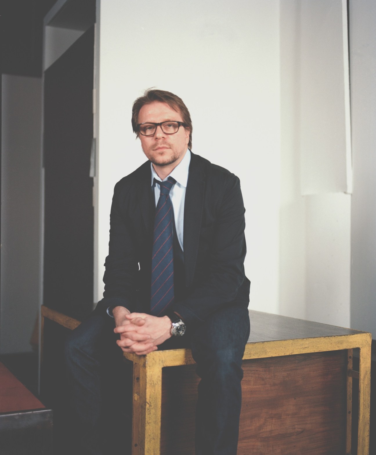 Man in dark blazer and striped tie leaning against wooden furniture in modern white interior.