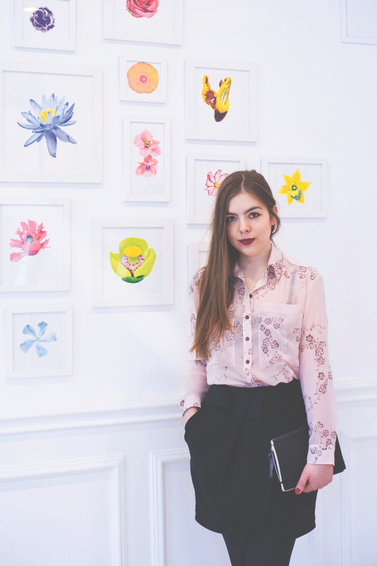 Woman standing in front of a white wall displaying framed watercolor flower illustrations in various colors.