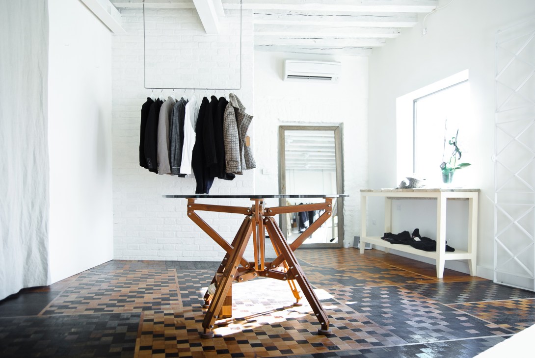 Interior of Laboratorio Barena boutique showing clothing on overhead rack, wooden display table, and shelving units in minimalist white space.