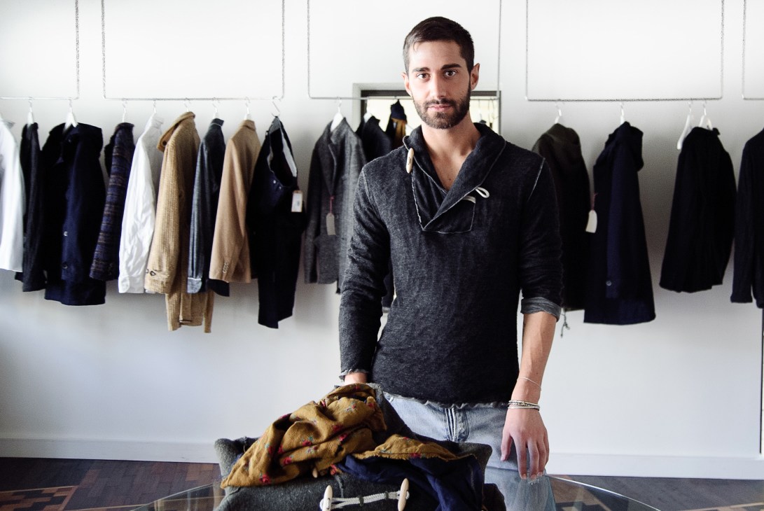 Man in grey shawl collar sweater stands before white wall with hanging clothing in various colors and styles.
