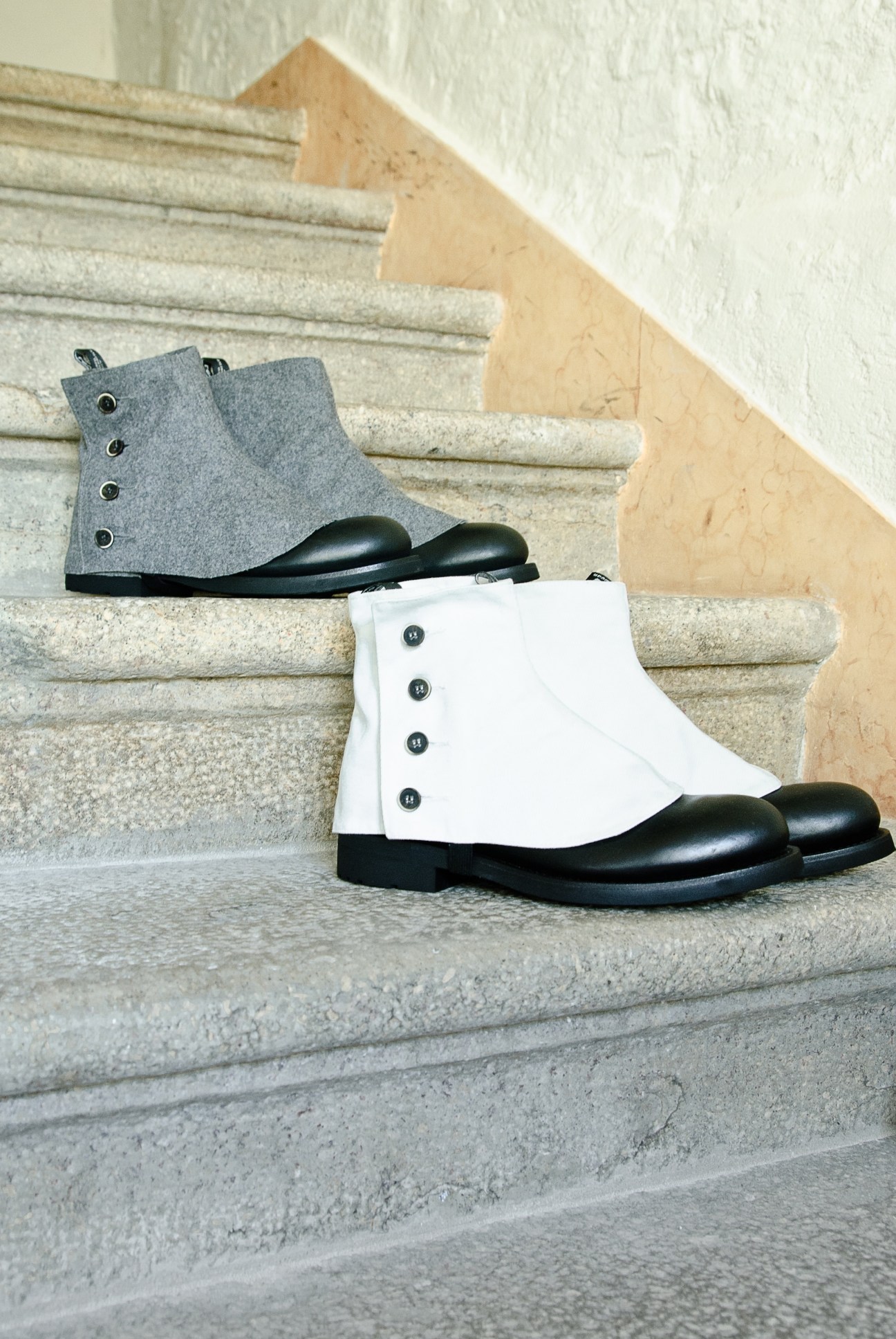 Two handmade leather ankle boots displayed on concrete steps, one grey and one white with black soles and button details.