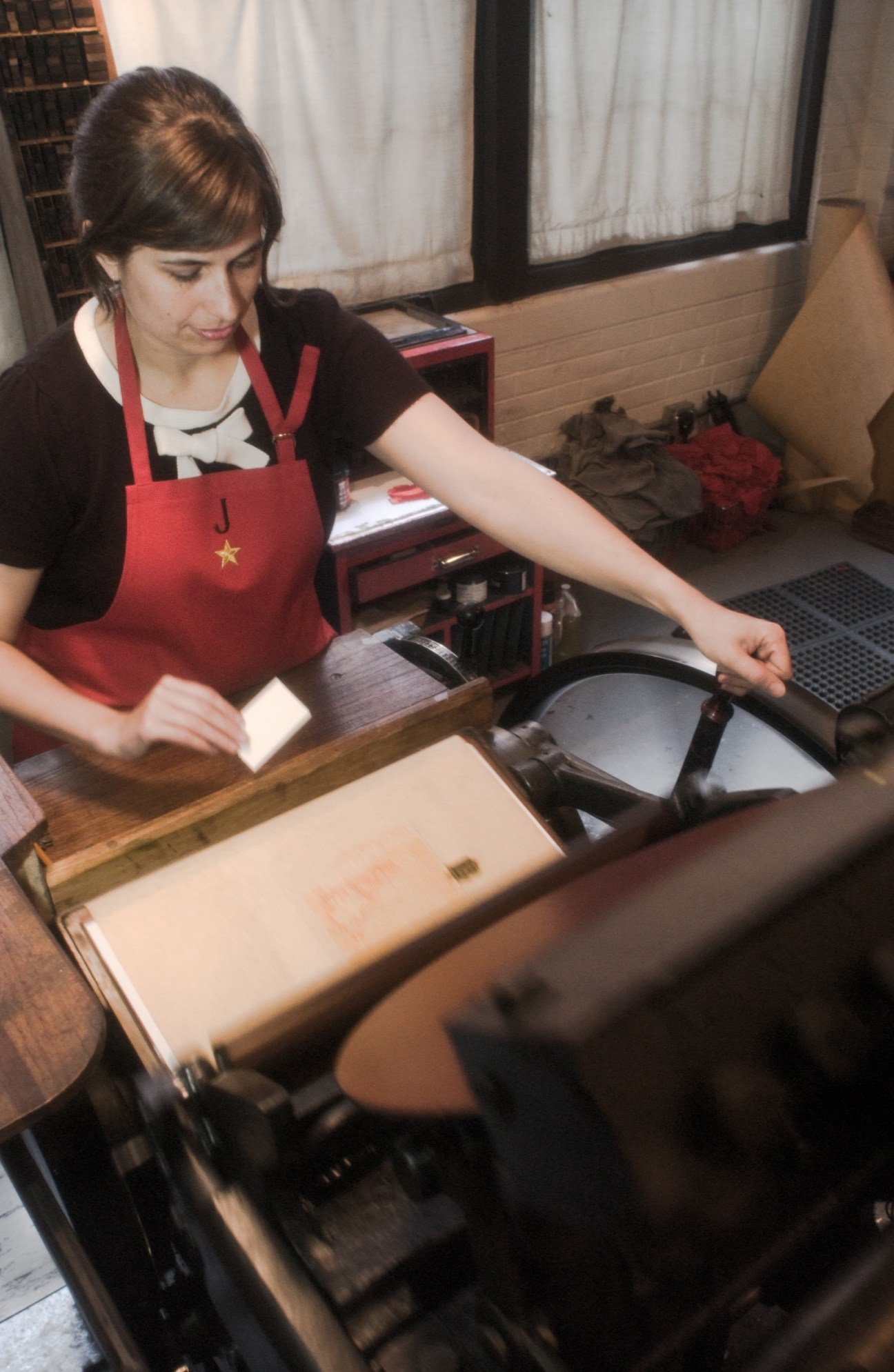 Woman in red apron operating an antique Chandler & Price letterpress printing machine.