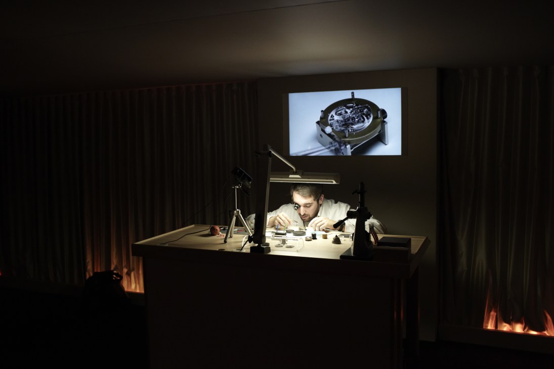 A watchmaker works at an illuminated desk with a screen displaying watch mechanics above.