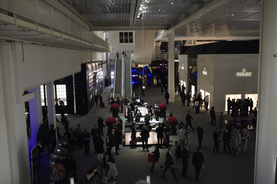 Aerial view of BaselWorld exhibition hall with multiple watch brand booths, visitors, and Rolex signage visible.
