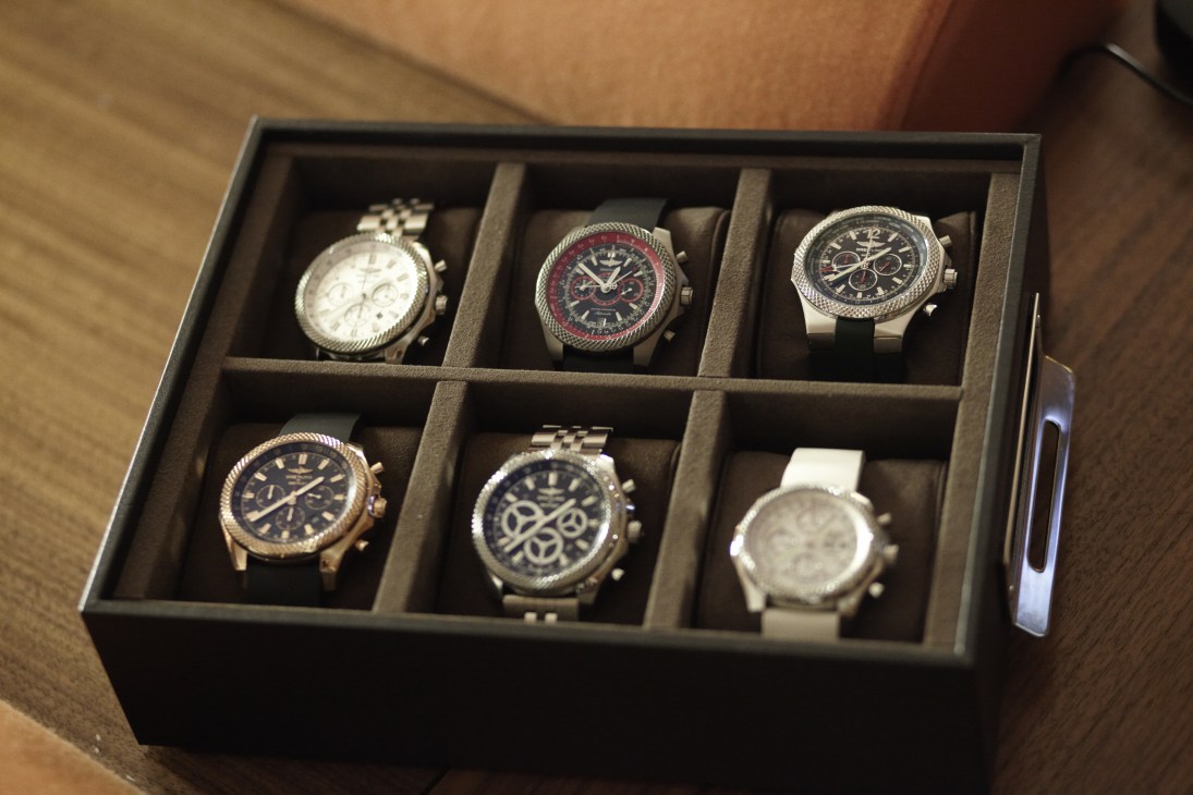 Six luxury chronograph watches displayed in a divided black presentation box with tan fabric lining.