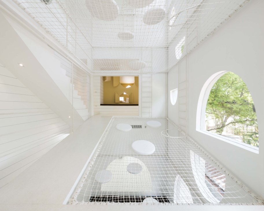 Minimalist white interior space with mesh ceiling, white walls, and circular window opening to greenery.