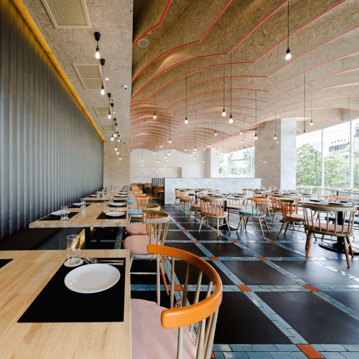 Modern restaurant interior with scallop-shaped plywood vaulted ceiling featuring neon-orange accents, wooden tables, orange chairs, and pendant lighting.