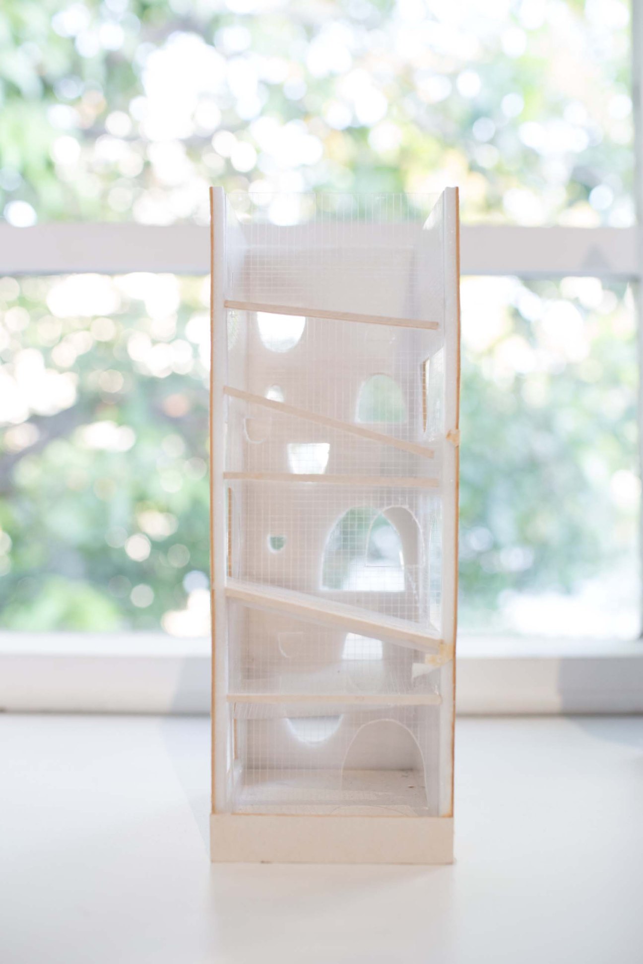 Wooden architectural scale model with translucent panels and cutout sections displayed against a blurred green background.