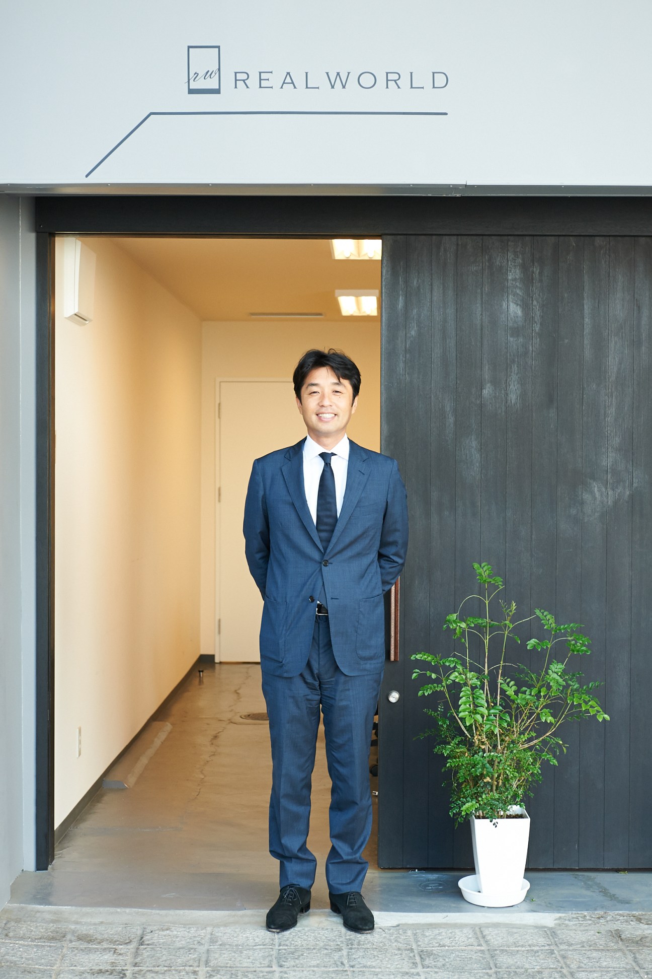 Masanao Idehara stands in the doorway of the Realworld office in Onomichi, wearing a blue suit and tie.