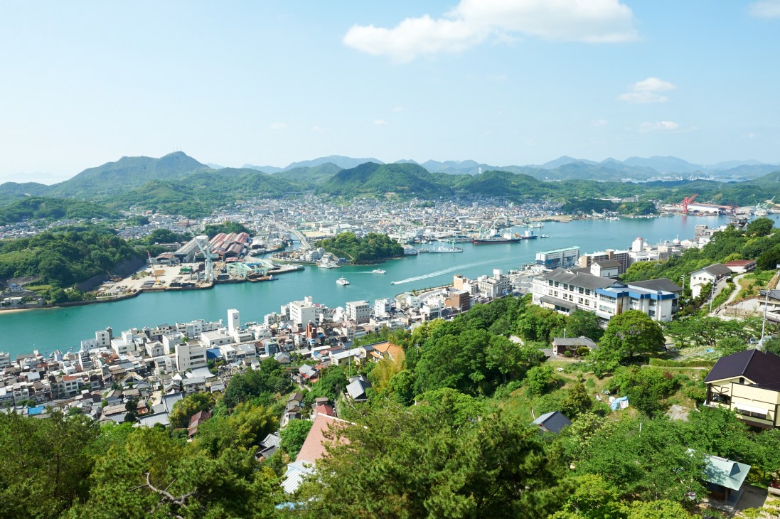 Aerial view of Onomichi city with turquoise waters of Seto Inland Sea, surrounded by green hills and mountains.