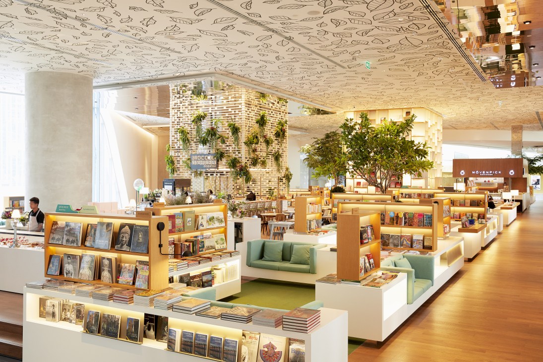 Interior of Open House bookstore with wooden shelving, green seating areas, plants, and cork ceiling at Central Embassy Bangkok.