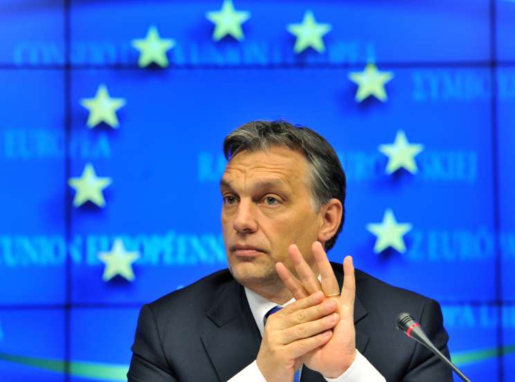 Man in dark suit speaking at microphone with EU flag backdrop featuring yellow stars.